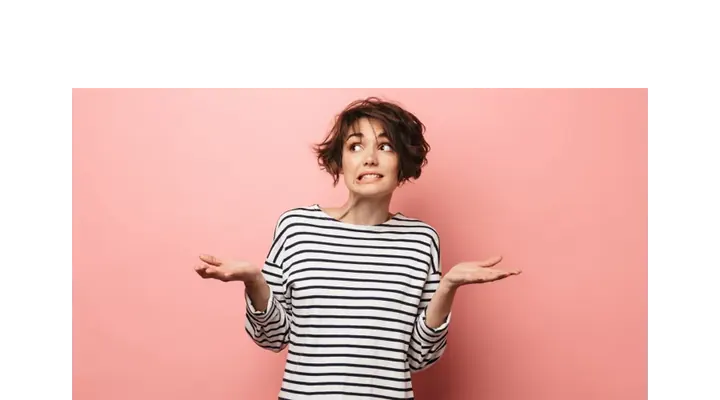 A female with short brown hair in a stripped black and white shirt. Standing in front of a pink background with her hands up looking confused.