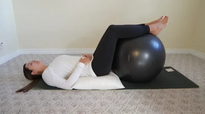 Laura, laying on her back on a black yoga mat. There is a white pillow under her pelvis and back. Her legs are up on a grey exercise ball. Her hands are resting on her pelvis. She is looking at the ceiling.