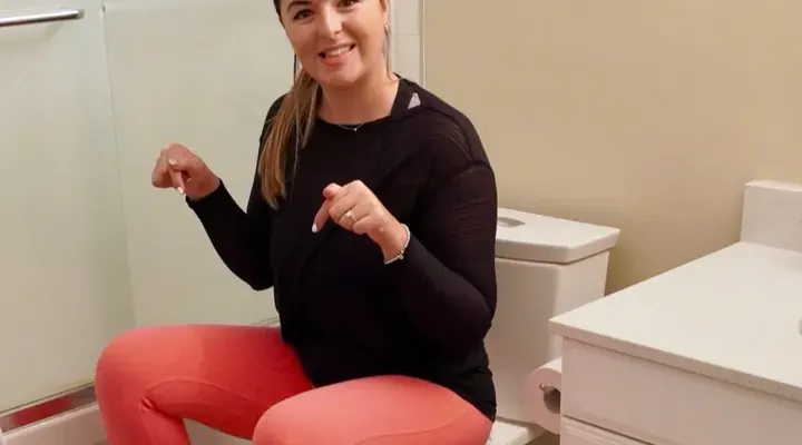 Laura in pink leggings and a black shirt. She is sitting on a white toilet with her feet on a white stool. She is point to the stool and smiling.